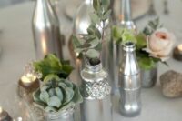 a chic and simple wedding centerpiece made of silver bottles and jars, with greenery, blooms and succulents plus candles around