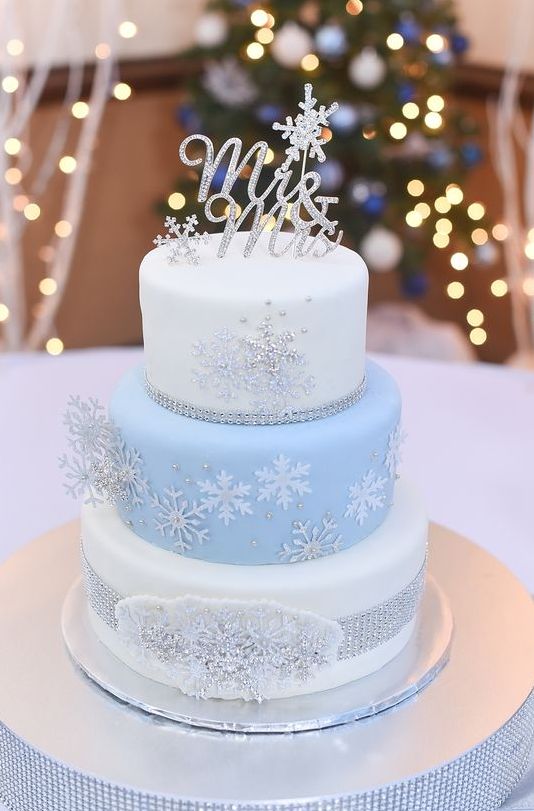 a glam white and light blue winter wedding cake decorated with silver and white snowflakes, topped with rhinestone ones and a calligraphy topper