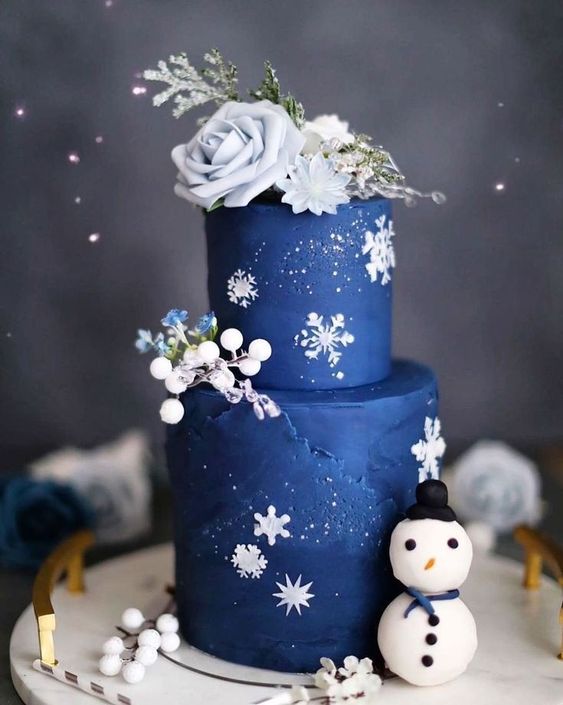 a navy wedding cake decorated with white snowflakes, frosted blooms on top, berries and blooms and a snowman by its side