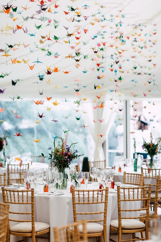 a neutral wedding reception space with bright blooms on the tables and colorful paper crane garlands over them