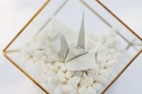a pretty neutral wedding decoration of a terrarium with pebbles and a grey paper crane is a subtle and cool idea
