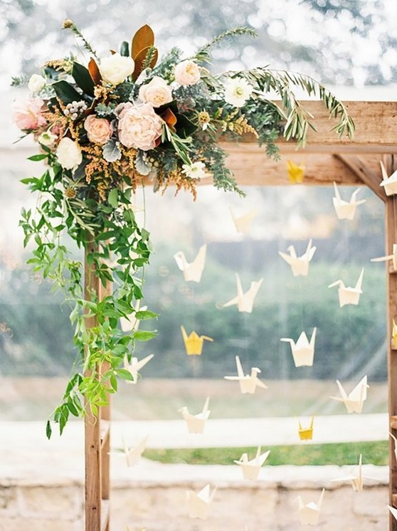 a rustic wedding arch with blush and neutral blooms and greenery and white and yellow origami cranes hanging down