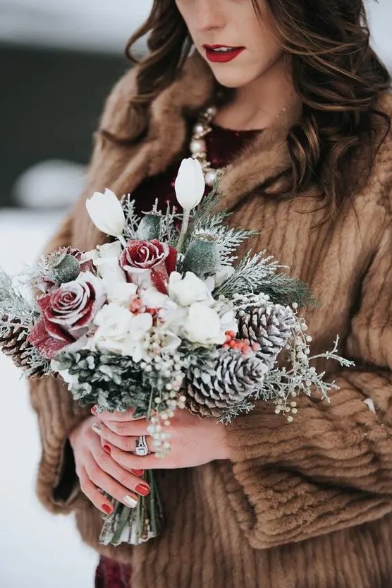 a snowy winter wedding bouquet of white blooms and snowy red roses, greenery, pinecones and seed pods is a gorgeous idea for winter