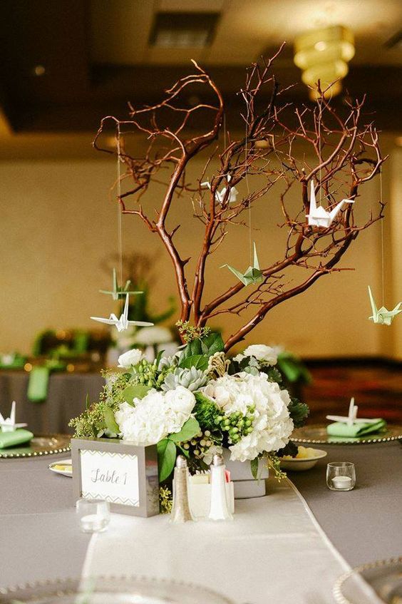a wedding centerpiece of white blooms, greenery and berries, with a tree with green and white paper cranes and some candles around
