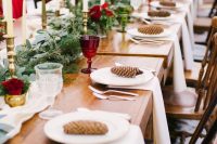 a winter wedding table setting with a fir and greenery runner, thin and tall candles, burgundy roses and glasses and pinecones