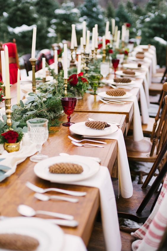 a winter wedding table setting with a fir and greenery runner, thin and tall candles, burgundy roses and glasses and pinecones