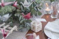 an ethereal wedding tablescape with a neutral table runner, lots of eucalyptus, candles, white plates and napkins andwhite and burgundy paper cranes