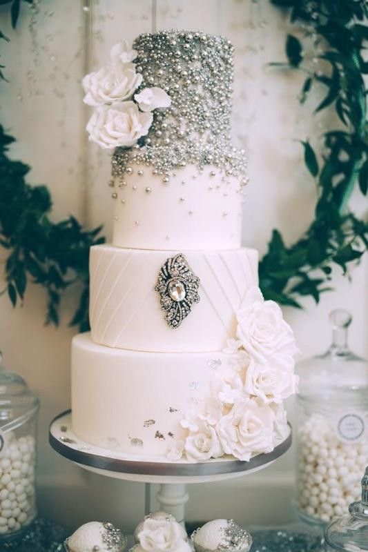 a beautiful white wedding cake with heavy embellishments and a large brooch, white blooms and silver foil for a glam wedding