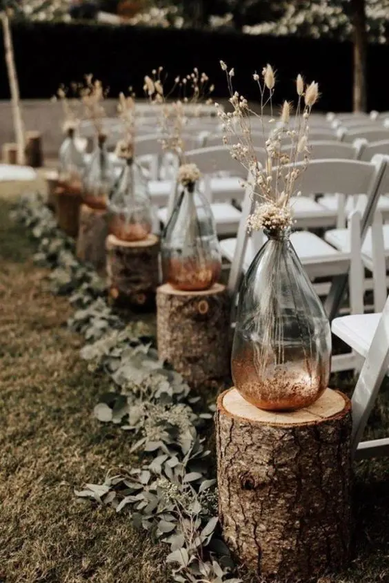 a boho wedding aisle with tree stumps, large irregular vases with dried grasses and a greenery runner is amazing