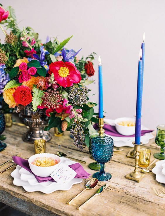 a bold Spanish-inspired wedding tablescape with hot pink, yellow and blue blooms, blue candles and glasses and purple napkins
