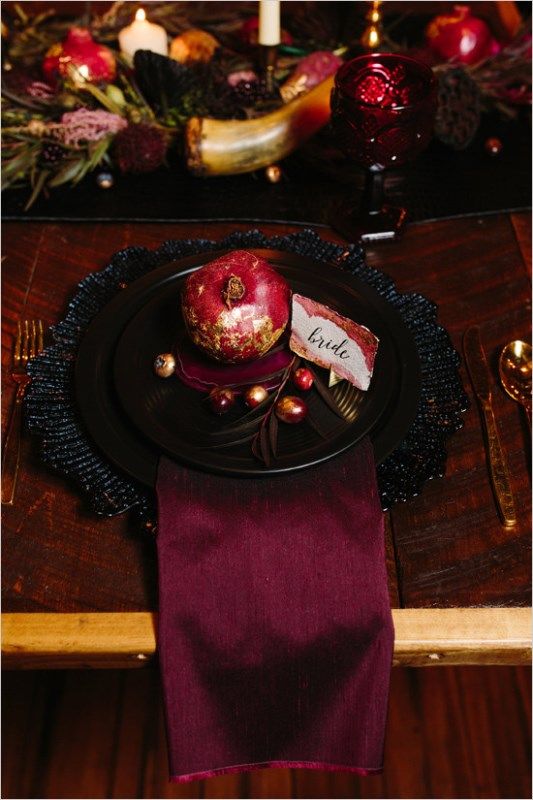 a bold place setting with black plates and a charger, a burgundy napkin and a gilded pomegranate