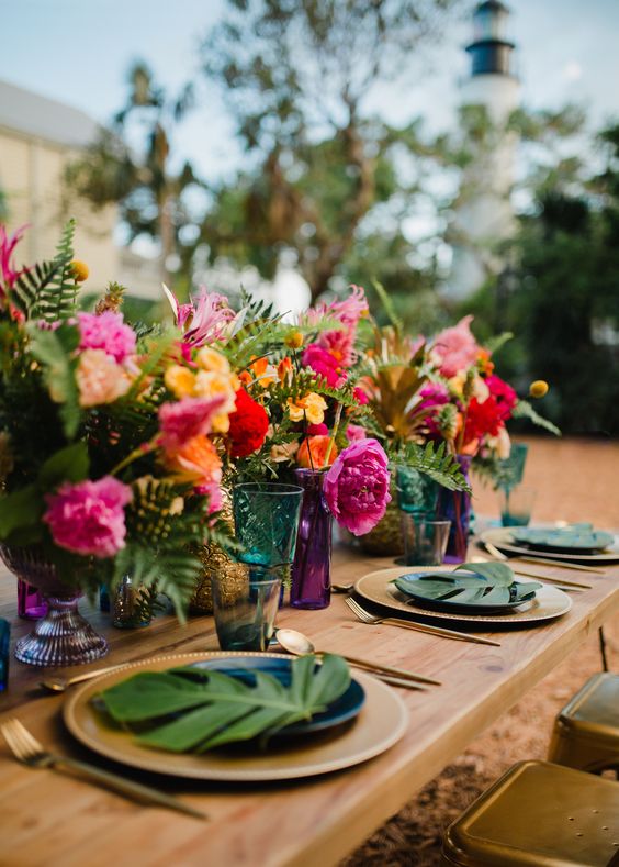 a bold tropical wedding table with bright florals and leaves, gold chargers and cutlery and colorful glasses and glass vases