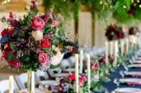 a bold wedding table setting with a navy tablecloth, fuchsia napkins, gold touches, pink, red and blue blooms and lush greenery chandeliers and a runner