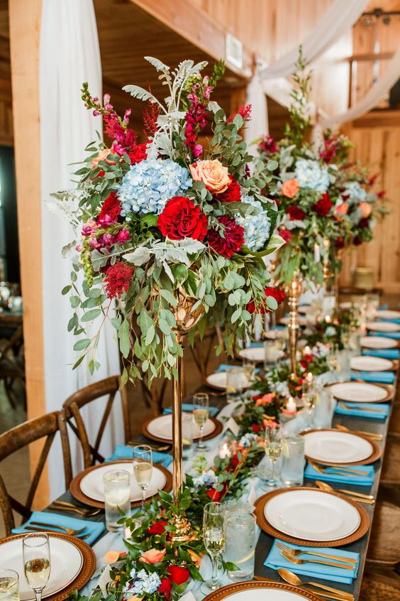 a bold wedding tablescape with a light blue table runner and napkins, woven placemats, deep red, blue and peachy blooms on tall stands