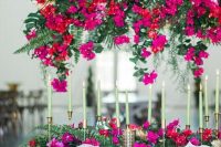 a bold wedding tablescape with hot pink, red blooms and greenery and tropical leaves, with gold touches and candlesticks plus mitn green candles