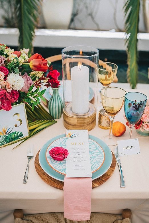 a bright and chic wedding tablescape with a blush tablecloth, pink napkins, yellow and blue glasses, red and pink blooms and greenery is a cool idea