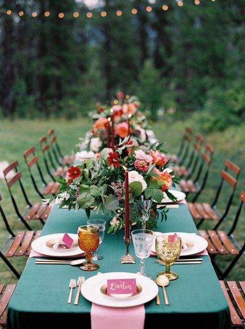 a bright wedding tablescape with a green tablecloth and pink napkins, red and orange blooms and greenery, amber glasses