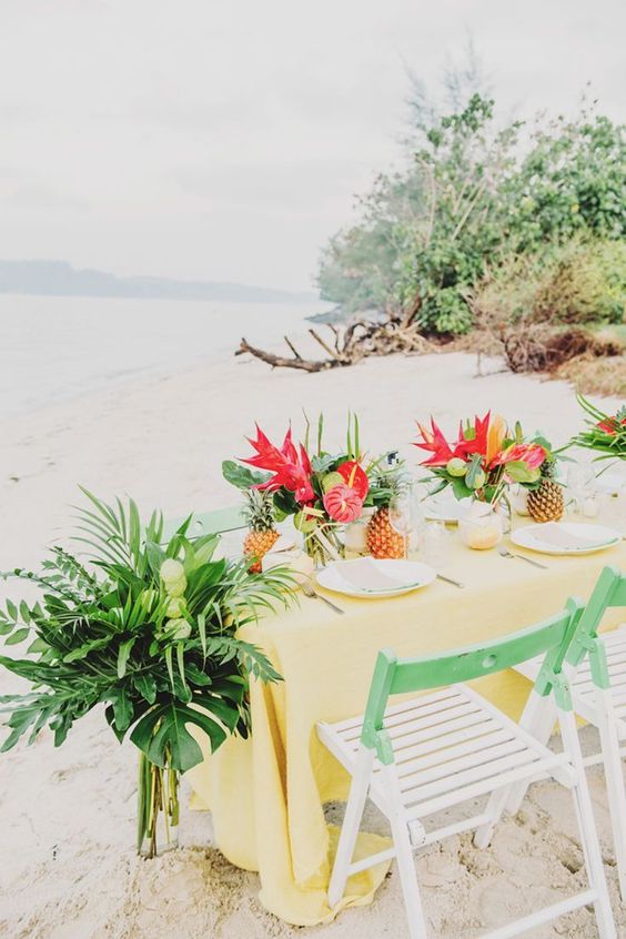 a bright wedding tablescape with a sunny yellow tablecloth, bold red blooms and greenery, tropical leaves and pineapples
