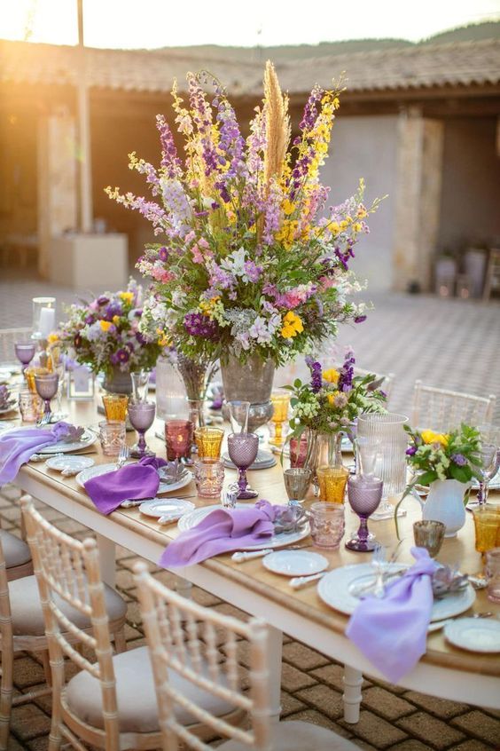 a cheerful summer wedding tablescape with lilac and purple napkins, lilac and amber glasses, yellow, lilac and purple blooms and greenery