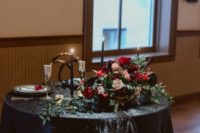 a chic head table with a black tablecloth, black candles in gold candle holders, a lush centerpiece with hot red and black blooms