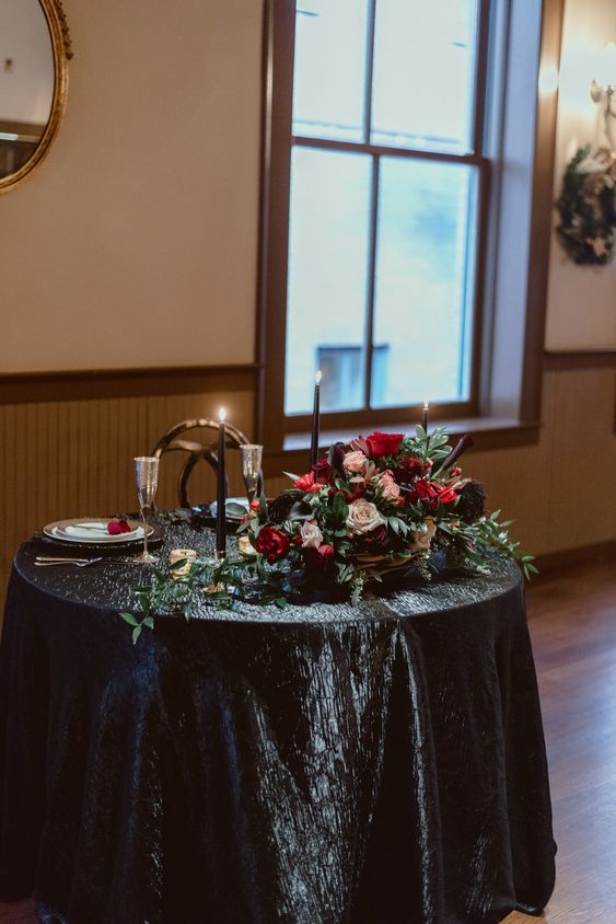 a chic head table with a black tablecloth, black candles in gold candle holders, a lush centerpiece with hot red and black blooms