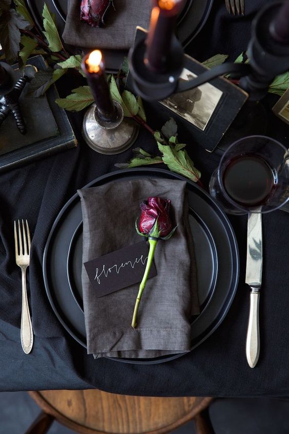 a chic modern black wedding table setting with matte plates, black candles, greenery and a single deep red rose