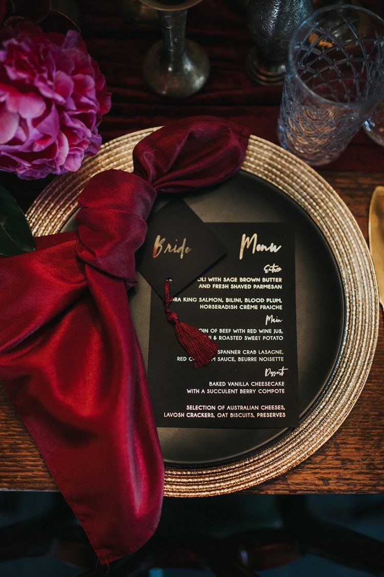 a chic place setting with a metallic charger, black plates and menus plus a deep red napkin