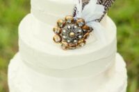 a chic white wedding cake with a lace tier and textural ones, with an oversized vintage brooch and feathers for a boho wedding