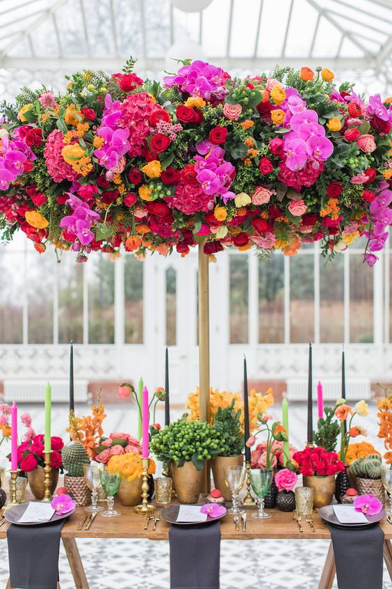 a colorful and luxurious wedding tablescape with fuchsia, hot pink, yellow and red blooms, greenery, colorful candles and an oversized floral installation over the table