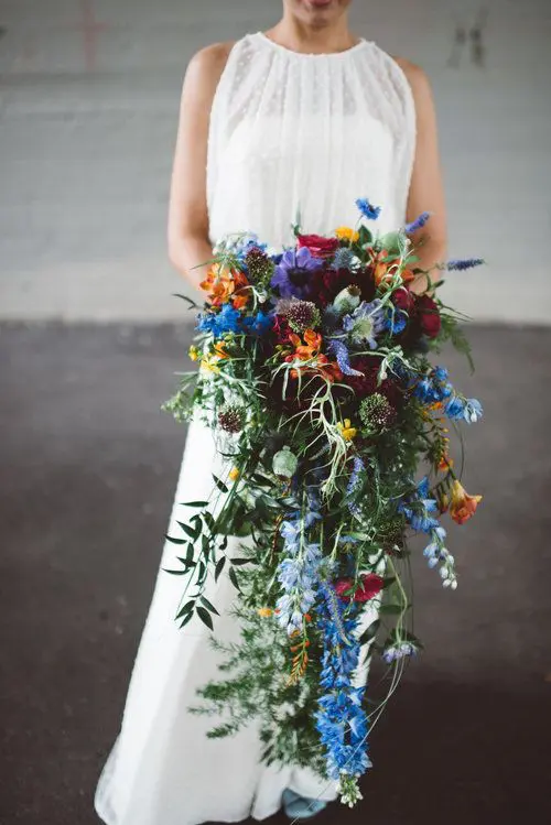 a colorful cascading wedding bouquet with blue, purple, burgundy and orange and yellow blooms and greenery is a gorgeous idea for a colorful wedding