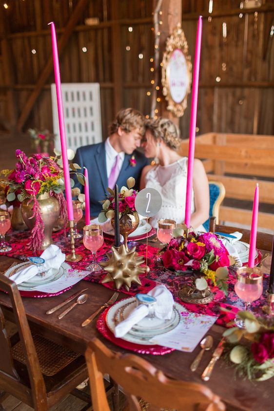 a colorful wedding tablescape with hot pink blooms, hot pink tall candles, hot pink chargers, greenery and bold and brass touches is cool