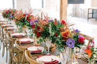 a colorful wedding tablescape with red napkins, red, pirple and orange blooms, greenery, feathers and fronds is a cool idea