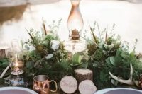 a gorgeous forest wedding tablescape with a lush greenery runner, antlers, stumps, lamps and copper touches