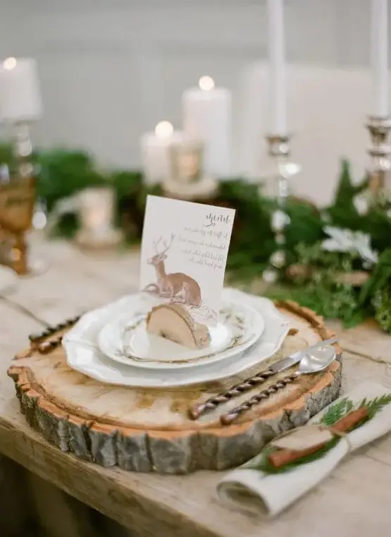 a gorgeous winter wedding tablescape with an evergreen runner and candles, a wood slic eplacemat and plates with a card