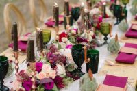 a jewel-tone wedding table setting with purple napkins and blooms, dark green candles and glasses, succulents