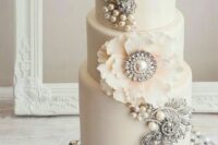 a neutral wedding cake decorated with pearls and brooches plus a large sugar bloom is a chic and stylish idea