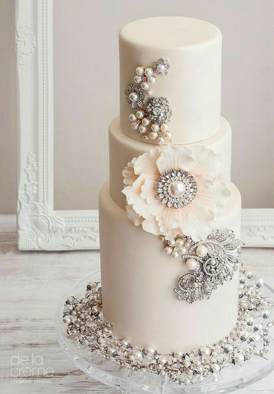 a neutral wedding cake decorated with pearls and brooches plus a large sugar bloom is a chic and stylish idea