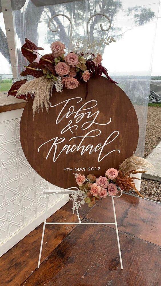 a pretty round wooden plaque sign with calligraphy, pink blooms and grasses is a cool idea for a boho glam wedding