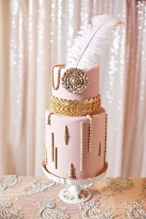 a refined art deco wedding cake in pink, with gold scallops, a large brooch and pearls plus a feather on top