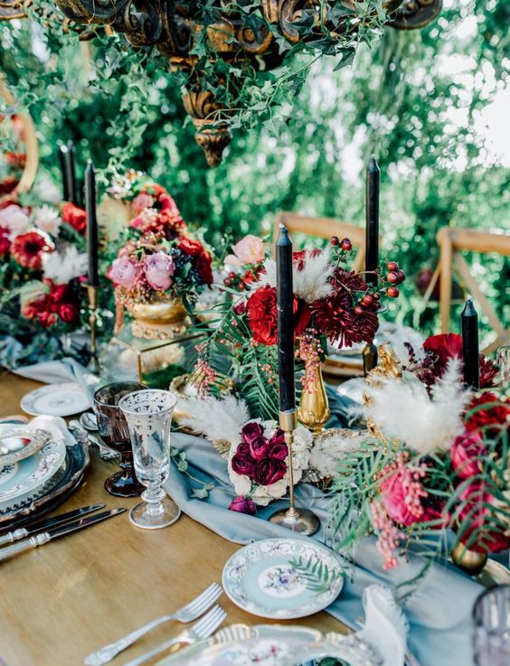 a refined secret garden wedding table setting with a blue table runner and floral plates, bold blooms and greenery, black candles and chic glasses