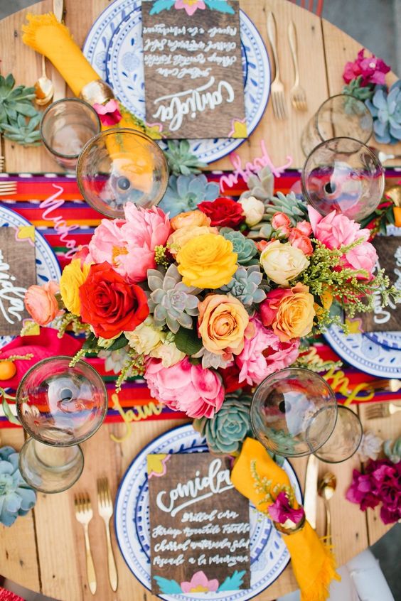 a vibrant wedding tablescape with red, pink, yellow blooms and succulents, a colorful table runner, plywood menus and yellow napkins
