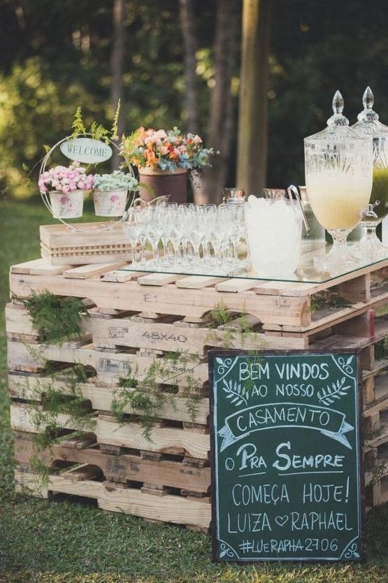 a wedding drink bar of pallets and ferns in between, with a chalkboard sign and floral arrangements on top