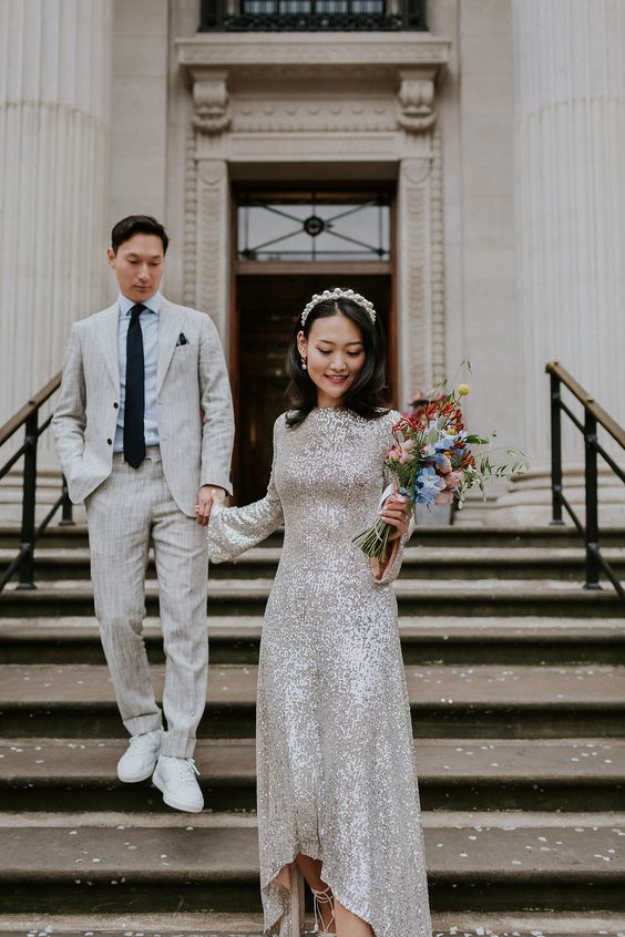 a white sequin high low wedding dress with bell sleeves and a high neckline, lace up shoes and a headband for a glam wedding