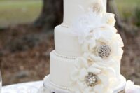 a white wedding cake with sugar blooms with brooches and feathers plus sugar cake toppers is a cool and bold idea