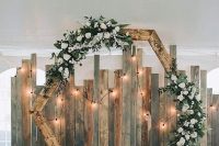 a wooden hexagon wedding arch with white blooms and greenery and string lights is a cool and lovely idea for a rustic wedding