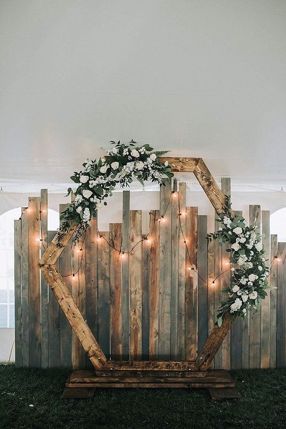 a wooden hexagon wedding arch with white blooms and greenery and string lights is a cool and lovely idea for a rustic wedding