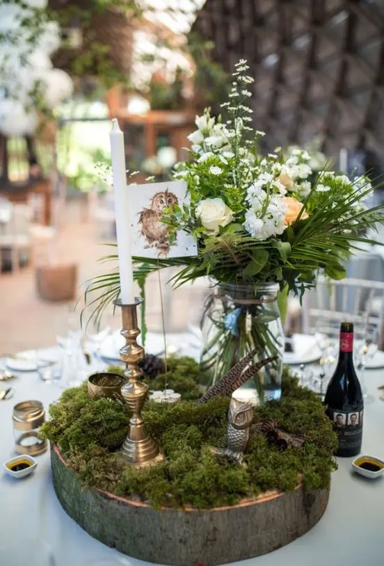 a woodland wedding centerpiece of a wood slice, moss, blooms and greenery in a jar, feathers and candles
