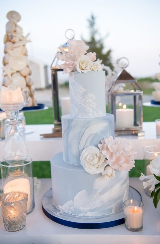 a blue watercolor wedding cake with sugar and real blooms and candles around for a beach wedding dessert table