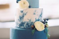 a chic modern blue wedding cake with a light blue, white watercolor and bold blue tier, white ranunculus, berries and gilded blackberries