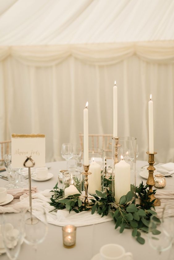 a chic wedding centerpiece of white table runners, greenery, various white candles and gold candleholders